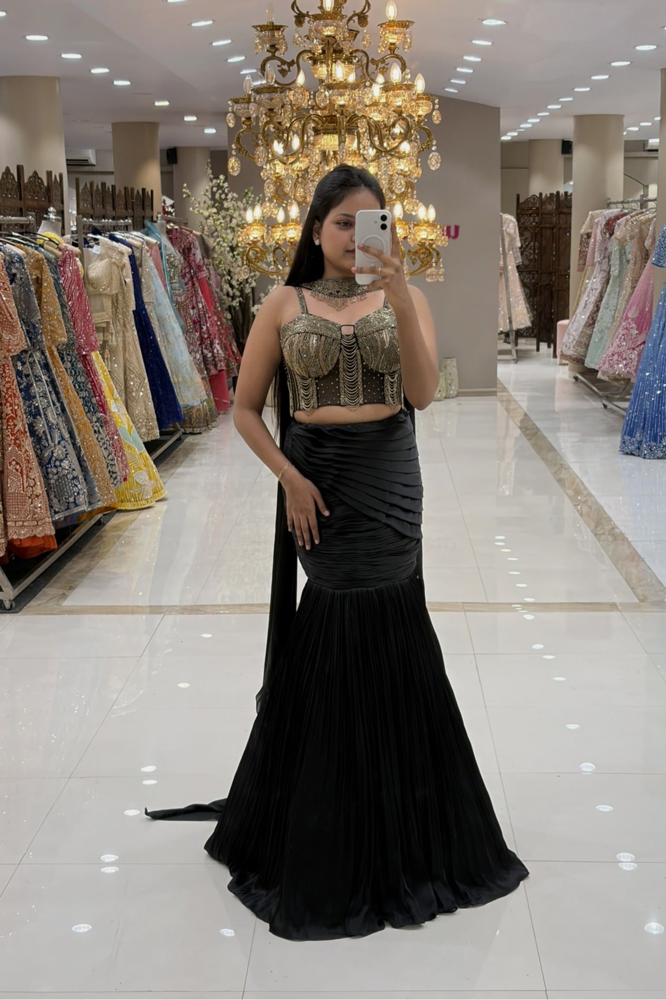 Woman taking a mirror selfie in a luxury Indo-Western black fishcut lehenga with an attached cape and a heavy gold handwork corset blouse at Urban Dulhaniya boutique.