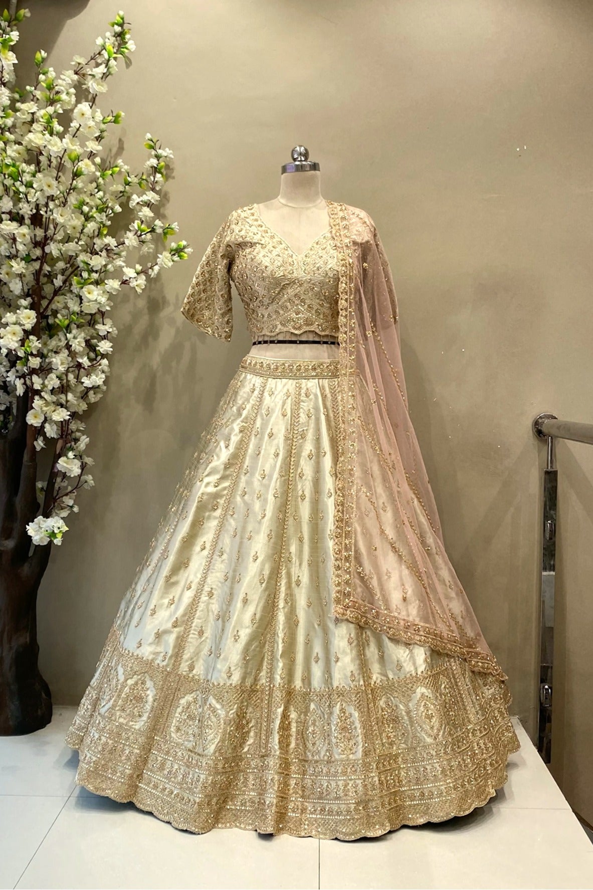 Full front view of a luxury ivory raw silk bridal lehenga with heavy gold zardosi embroidery and a peach scalloped dupatta at Urban Dulhaniya Pune.