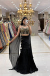 Woman taking a mirror selfie in a luxury Indo-Western black fishcut lehenga with an attached cape and a heavy gold handwork corset blouse at Urban Dulhaniya boutique.
