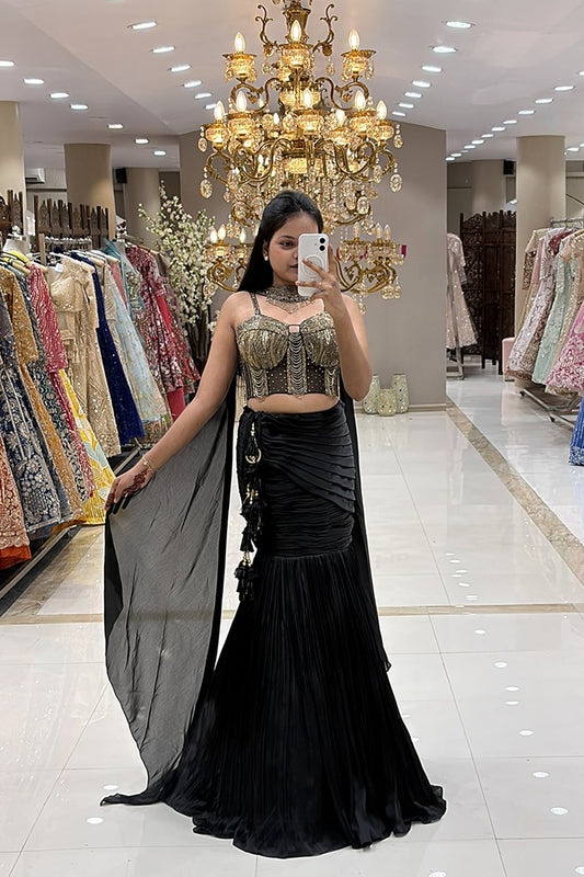 Woman taking a mirror selfie in a luxury Indo-Western black fishcut lehenga with an attached cape and a heavy gold handwork corset blouse at Urban Dulhaniya boutique.
