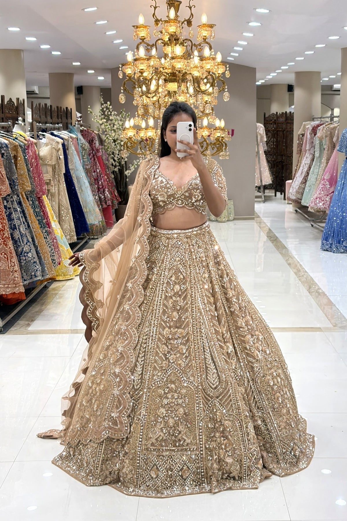Full-length mirror selfie of a bride wearing a luxury champagne gold heavy handwork bridal lehenga with a scalloped dupatta at the Urban Dulhaniya boutique.