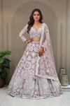 Mauve lavender designer lehenga with ivory pearlwork and cutdana floral embroidery, featuring a V-neck blouse and scalloped net dupatta against a fluted wall background.