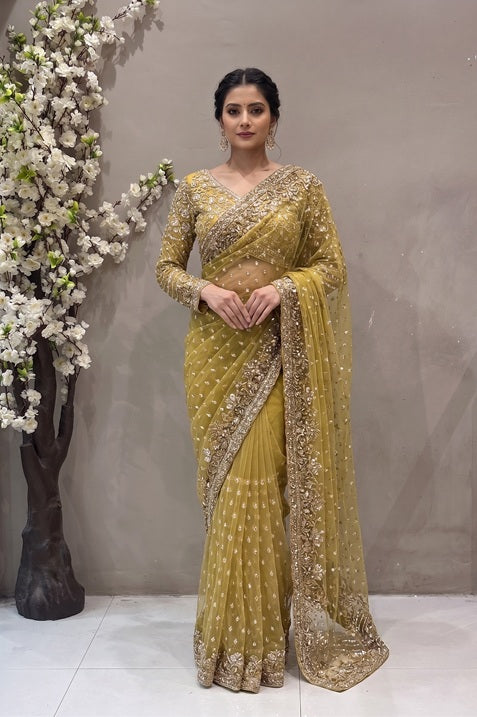 Model wearing a mustard gold net saree with heavy zardosi floral embroidery border and a full-sleeved embellished blouse against a neutral background.
