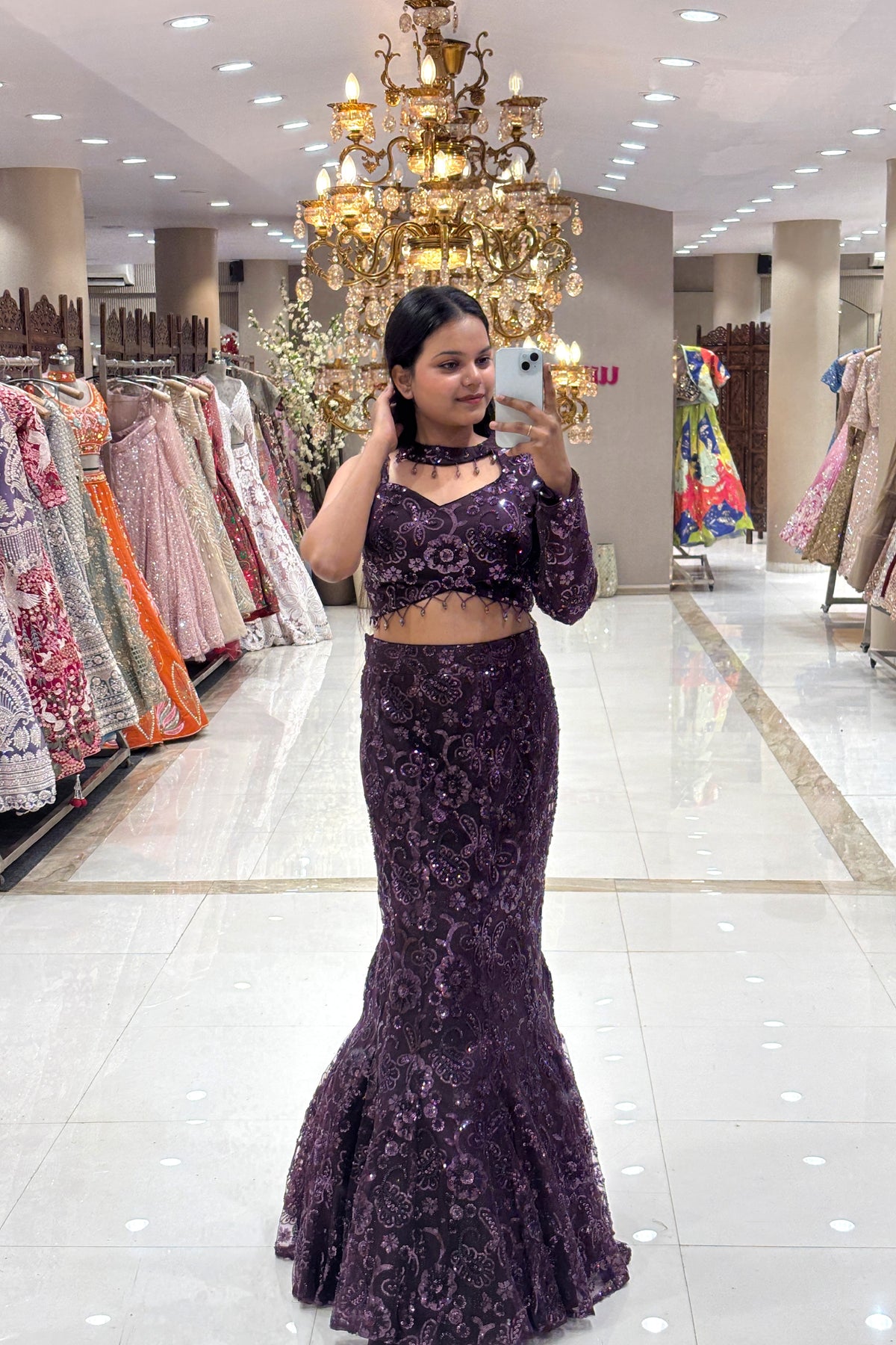 Full-length view of a woman taking a mirror selfie wearing a luxury plum purple fishcut Indo-Western lehenga with an asymmetrical one-shoulder blouse at the Urban Dulhaniya boutique.